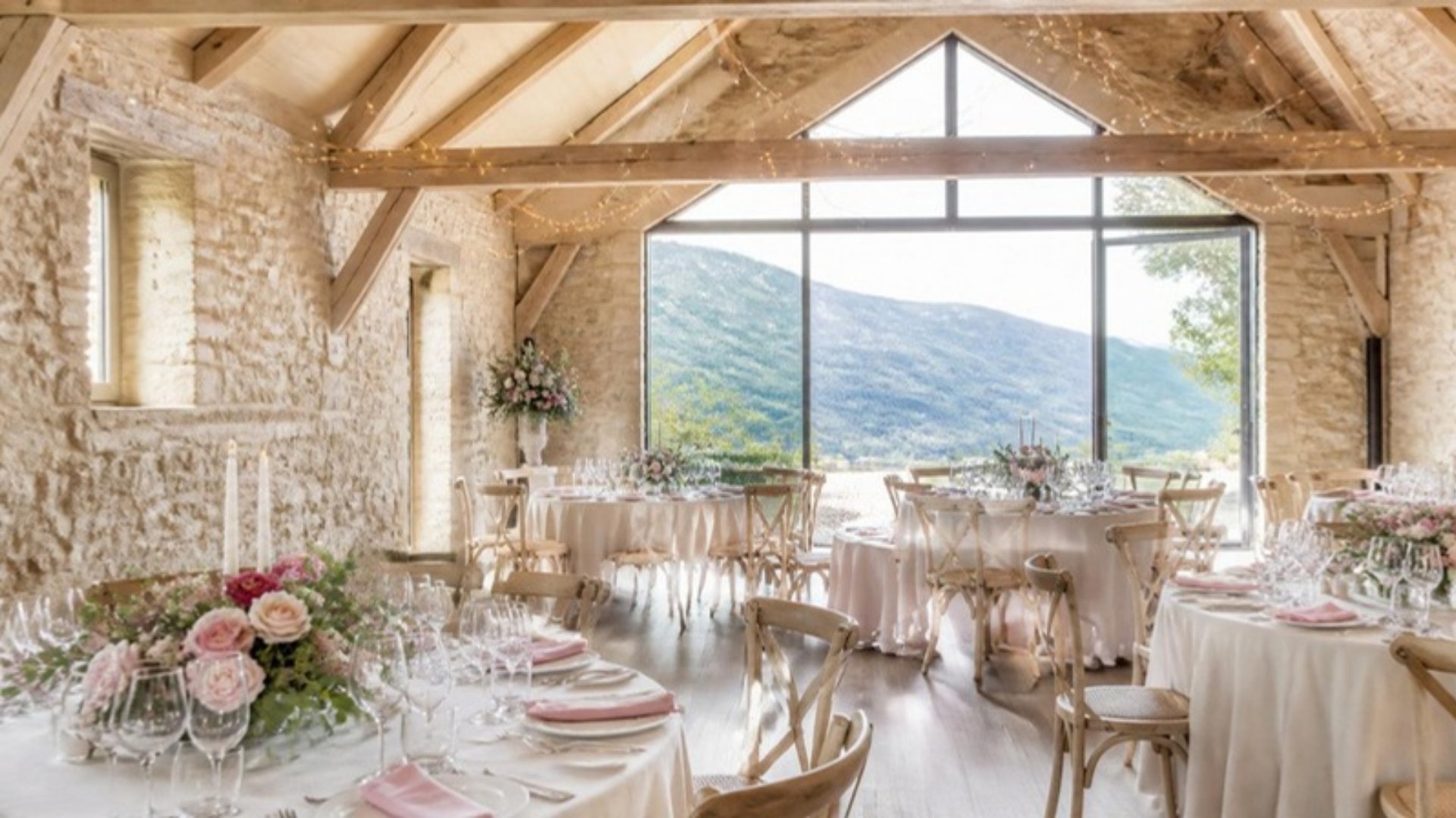 Salle de réception en pierre et poutres dressée pour un mariage, baie vitrée ouverte sur les montagnes